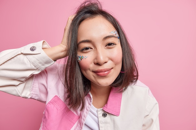 Nahaufnahme eines schönen asiatischen tausendjährigen Mädchens mit dunklem Haar, natürliches Make-up, gesunde Haut hat glitzernde Steine auf dem Gesicht, gekleidet in modische Jacke, isoliert über rosa Wandblicken zufrieden