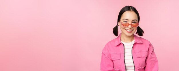 Nahaufnahme eines schönen asiatischen weiblichen Modells mit stylischer Sonnenbrille, das vor rosafarbenem Hintergrund im trendigen Outfit-Kopienraum posiert