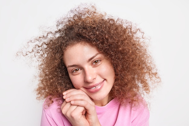 Nahaufnahme eines schönen jungen weiblichen Modells hält die Hände in der Nähe des Gesichts neigt den Kopf hat natürliches lockiges Haar, gesunde Haut bewundert etwas Schönes, isoliert über weißer Wand. Menschliche Gesichtsausdrücke