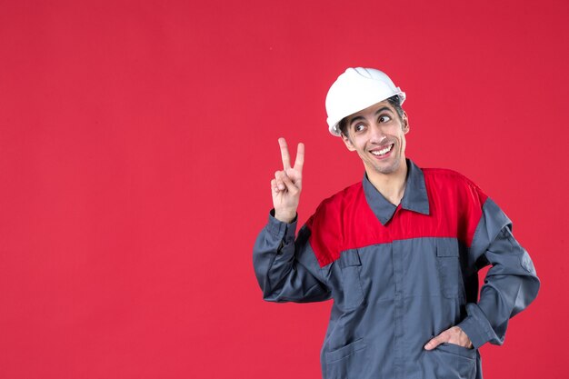 Nahaufnahme eines selbstbewussten jungen Architekten in Uniform mit Schutzhelm und Siegesgeste auf isolierter roter Wand