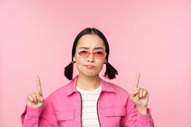 Kostenloses Foto nahaufnahme eines stilvollen koreanischen mädchens, das ratlos aussieht und mit dem finger nach oben zeigt und werbung zeigt, die über rosa hintergrund steht