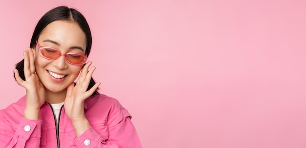 Nahaufnahme eines stilvollen koreanischen Mädchens mit Sonnenbrille, das glücklich lächelt und vor rosafarbenem Hintergrund posiert