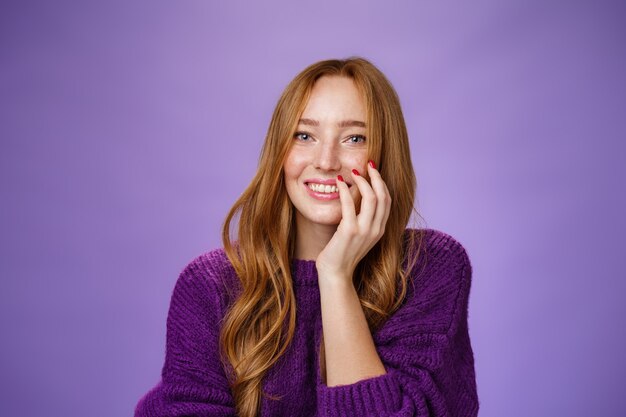 Nahaufnahme eines weiblichen und glamourösen, süßen Rothaarigen mit Sommersprossen in lila Strickpullover, der die Hand auf der Wange hält, sanft kichert und breit in die Kamera über violettem Hintergrund lächelt.