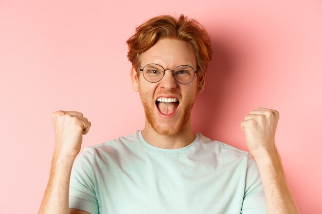 Nahaufnahme eines zufriedenen, glücklichen Rothaarigen, der gewinnt, vor Freude schreit und Faustpumpe macht, feiert den Sieg und steht wie ein Champion auf rosafarbenem Hintergrund.