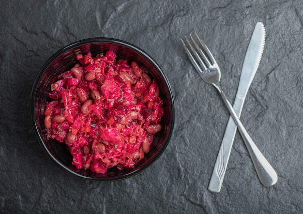 Nahaufnahme Foto von hausgemachter Vinaigrette mit Bohnen in schwarzer Schüssel mit Gabel und Messer auf schwarzem Hintergrund.