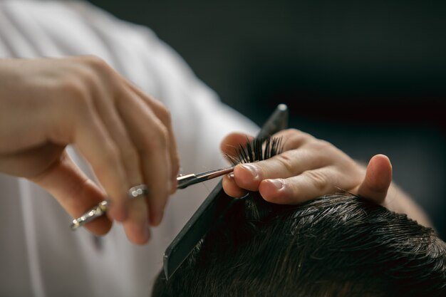 Nahaufnahme Hände des Friseurmeisters, Stylist mit Sccissors macht die Frisur zu Kerl, junger Mann. Beruflicher Beruf, männliches Schönheitskonzept. Haarpflege des Kunden. Weiche Farben und Fokus, Vintage.