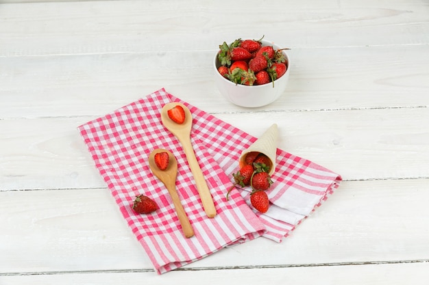 Nahaufnahme Holzlöffel und ein Kegel Erdbeeren auf roter Gingham-Tischdecke mit einer Schüssel Erdbeeren auf weißer Holzbrettoberfläche. horizontal