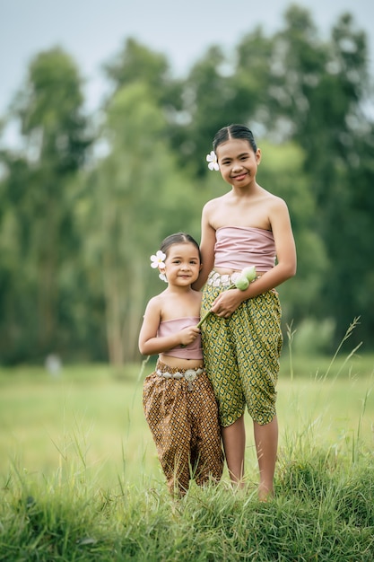 Nahaufnahme, Porträt der süßen Schwester und der jungen Schwester in thailändischer Tracht und weiße Blume auf ihr Ohr legen, die im Reisfeld steht, Lächeln, Geschwisterliebekonzept, Kopienraum