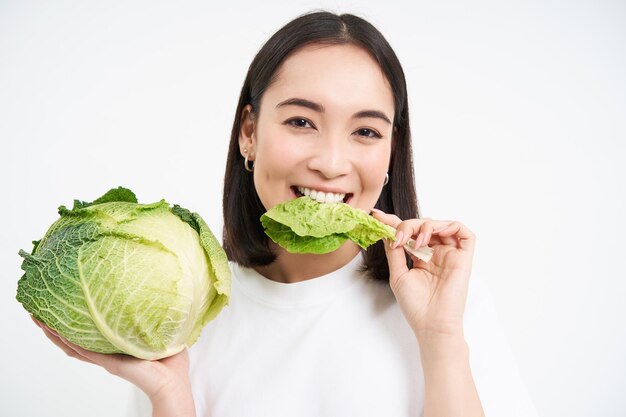 Nahaufnahme Porträt einer asiatischen Frau, die Salat beißt, Grünkohl isst und vor weißem Hintergrund lächelt