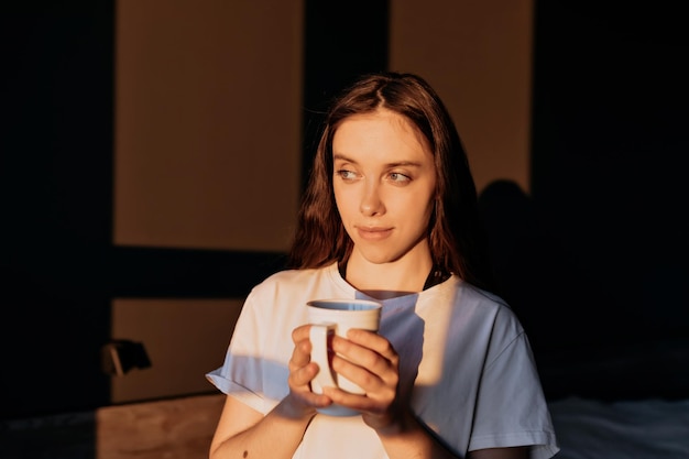 Nahaufnahme Porträt einer charmanten Dame mit dunklem Haar in weißem T-Shirt hält eine Tasse und blickt nach dem Aufwachen am Morgen bei Sonnenschein auf das Fenster