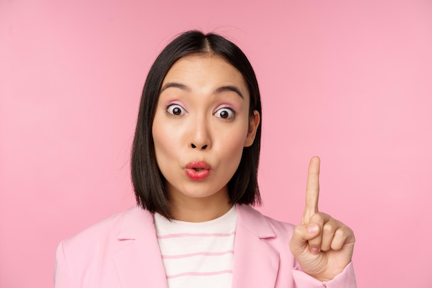 Nahaufnahme Porträt einer Geschäftsfrau, die den Finger nach oben hebt und vorschlägt, eine Idee oder Lösung zu haben, die über rosa Hintergrund steht