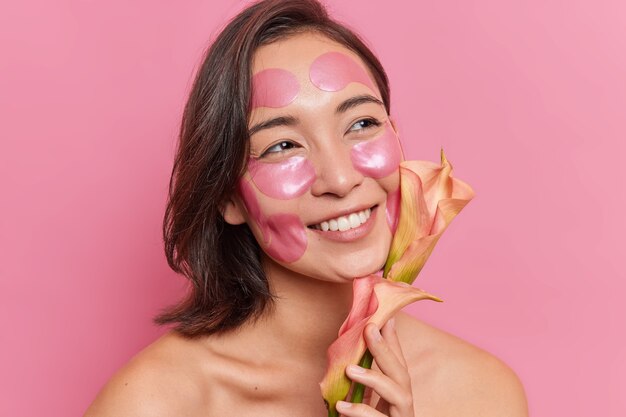 Nahaufnahme Porträt einer glücklichen jungen asiatischen Frau mit einem zahnigen Lächeln, die froh ist, eine Blume zu bekommen, trägt Hydrogel-Patches auf das Gesicht auf, um die Haut zu erfrischen, steht ohne Hemd an der rosa Wand