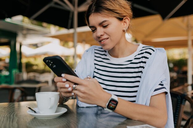 Nahaufnahme Porträt einer lächelnden Europäerin in gestreiftem T-Shirt in Smartwatch benutzt Smartphone und trinkt Morgenkaffee im Café im Freien