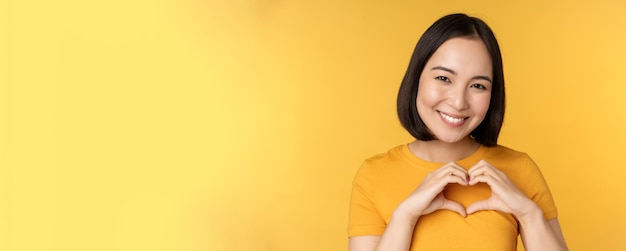 Nahaufnahme Porträt einer lächelnden Koreanerin, die ein romantisches Herzzeichen zeigt und glücklich über gelbem Hintergrund steht