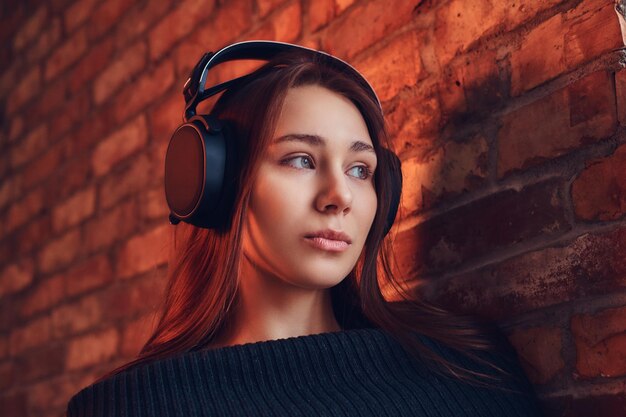 Nahaufnahme Porträt einer schönen charmanten Brünette in schwarzem Pullover hört Musik über gute Kopfhörer, während sie sich in einem Raum mit Loft-Interieur an eine Ziegelwand lehnt.