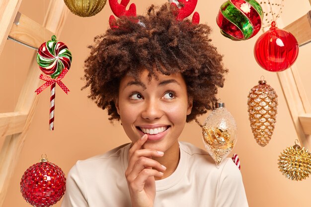 Nahaufnahme Schuss der attraktiven positiven jungen Frau hat breites Lächeln weiße Zähne lockiges buschiges Haar gekleidet in Freizeitkleidung Träume über Wunder am Neujahrstag umgeben von Weihnachtsspielzeug über Kopf