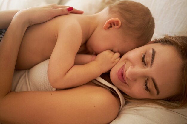 Nahaufnahme Schuss der glücklichen charmanten jungen Mutter, die mit ihrem schläfrigen kleinen Kind auf der Brust auf dem Bett liegt. Hübsche Mutter und Kind kuscheln im Schlafzimmer. Konzept für Liebe, Glück, Mutterschaft und Elternschaft