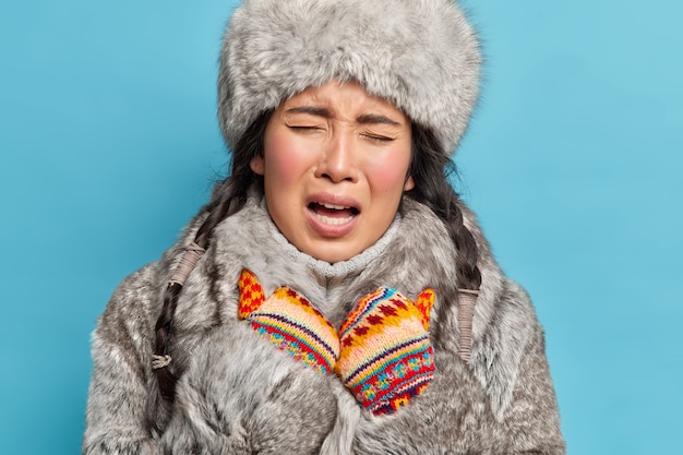 Kostenloses Foto nahaufnahme schuss der unzufriedenen jungen asiatischen inuitfrau trägt gestrickte handschuhe und winteroutfit fühlt sich kalt während des frostigen winters isoliert über blauer wand