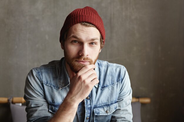 Nahaufnahme Schuss des attraktiven gut aussehenden jungen europäischen bärtigen Mannes gekleidet in modischem Jeanshemd und kastanienbraunem Hut lächelnd, nachdenklich, tief und weise Blick, seinen Bart berührend