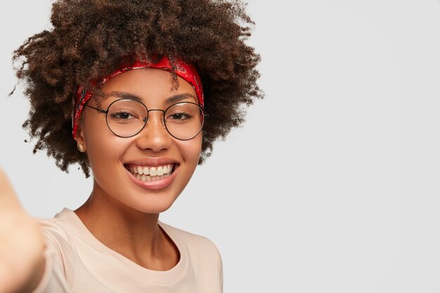 Nahaufnahme Schuss froh, dass weibliches Mädchen zahniges Lächeln hat, Afro-Frisur, schöne saubere Haut, trägt transparente Brille, streckt Hand als hält nicht erkennbares Gerät, macht Selfie über weiße Wand