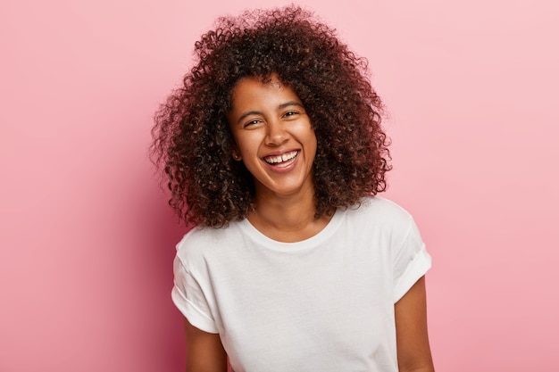 Nahaufnahme Schuss von hübschem Teenager-Mädchen mit dunkler Haut, lockigem Afro-Haar, grinst, hat weiße Zähne, lacht aufrichtig über guten Witz, hat Spaß mit engen Freund, trägt jeden Tag weißes T-Shirt