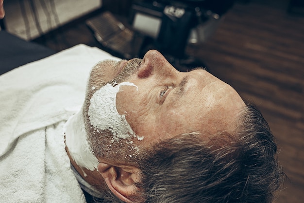 Nahaufnahme seitlicher Draufsicht hübscher älterer bärtiger kaukasischer Mann, der Bartpflege im modernen Friseursalon erhält.