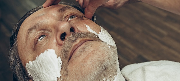 Nahaufnahme seitlicher Draufsicht hübscher älterer bärtiger kaukasischer Mann, der Bartpflege im modernen Friseursalon erhält.