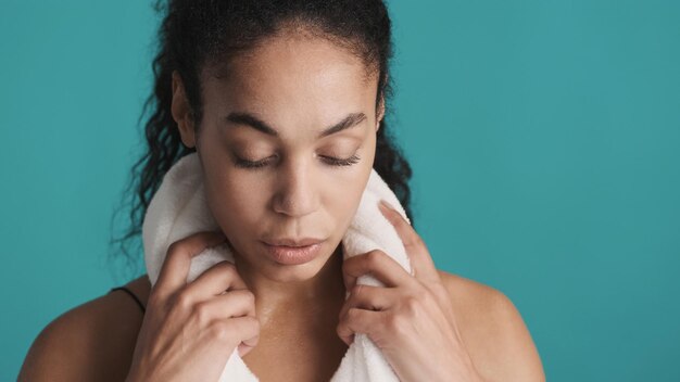 Nahaufnahme sportliche Afro-Frau, die ein Handtuch auf den Schultern hält und nach dem harten Training über blauem Hintergrund müde aussieht Mädchen beim Training