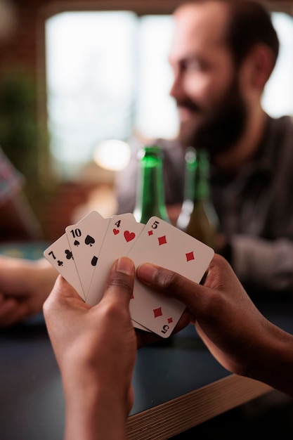 Nahaufnahme von afroamerikanischen Händen, die Karten halten, während sie zu Hause mit Freunden spielen. Verschiedene Gruppen von Menschen sitzen zu Hause im Wohnzimmer und entspannen sich bei Snacks, Bier und Gesellschaftsspielen.