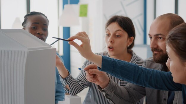 Nahaufnahme von Arbeitskollegen, die Gebäudemodell und Maquette auf dem Tisch analysieren, um die Konstruktion zu entwerfen. Gruppe verschiedener Kollegen, die an Architekturprojekten für Struktur und Entwicklung arbeiten.