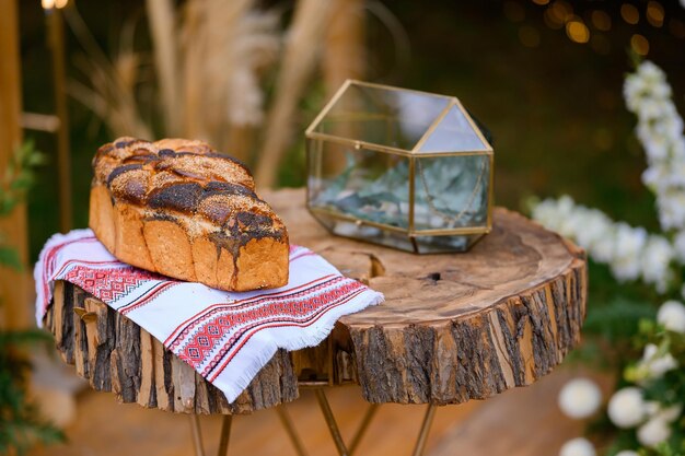 Nahaufnahme von gebackenem Brot auf einem Stickhandtuch, das auf einem Holztisch in der Nähe einer Glasbox platziert wurde, die für die Hochzeitszeremonie im Freien vorbereitet wurde