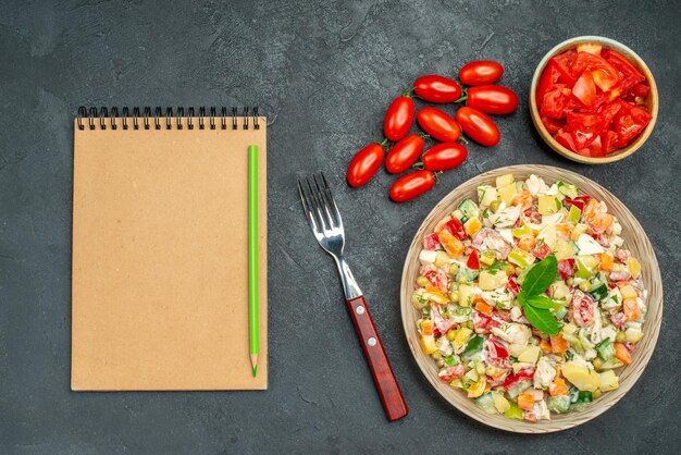 Nahaufnahme von Gemüsesalat mit Tomatengabel und Notizblock auf der Seite auf dunkelgrauem Hintergrund