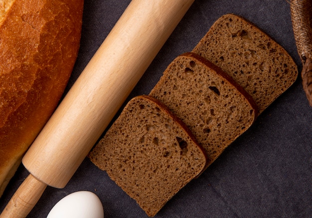 Nahaufnahme von geschnittenem roggenbrot und nudelholz mit ei auf kastanienbraunem hintergrund