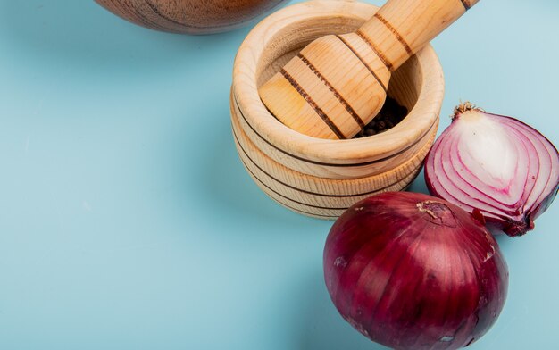 Nahaufnahme von geschnittenen und ganzen roten Zwiebeln und schwarzen Pfeffersamen im Knoblauchbrecher auf blauem Hintergrund