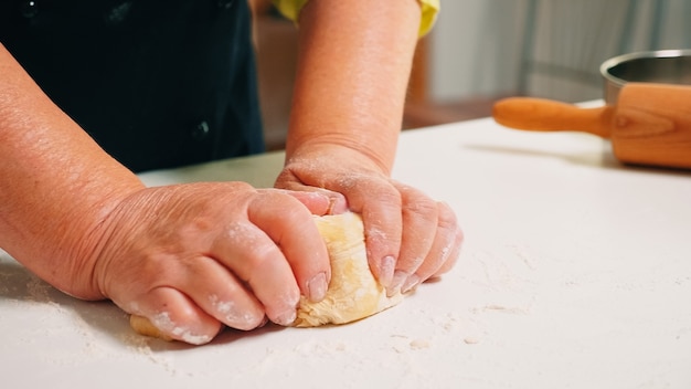 Nahaufnahme von Großmutterhänden, die auf dem Tisch in der heimischen Küche kneten. Pensionierter älterer Bäcker mit Bonete, der Zutaten mit gesiebtem Weizenmehl knetet, um traditionellen Kuchen und Brot zu backen.