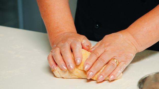 Nahaufnahme von Händen, die ein großes Stück Gebäck nehmen und Brötchen aus Teig machen. Pensionierter älterer Bäcker mit Bonete, der Zutaten mit gesiebtem Weizenmehl knetet, um traditionellen Kuchen und Brot zu backen.