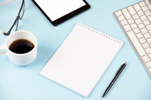 Nahaufnahme von Kaffee; Brille; digitales Tablett; Tastatur; unbelegter gewundener Notizblock und Stift gegen blauen Hintergrund