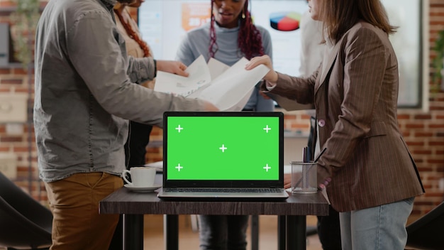 Nahaufnahme von Kollegen, die einen Laptop mit grünem Bildschirm auf dem Schreibtisch haben, mit Dokumenten arbeiten und einen isolierten Chroma-Key-Kopierbereich auf dem Computer haben. Arbeitskollegen, die eine leere Mock-up-Vorlage und einen Hintergrund verwenden.