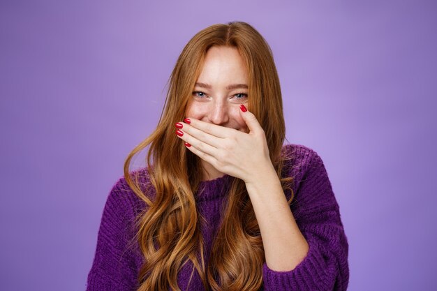 Nahaufnahme von Mädchen, die Spaß am Kichern haben und den Mund mit der Handfläche bedecken, während sie lachen, aufrichtig und sorglos lächeln und auf urkomische Witze oder Streiche reagieren, die auf violettem Hintergrund posieren.