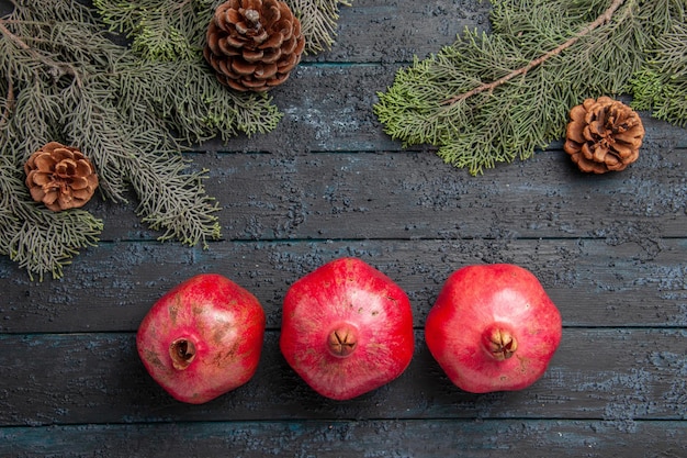 Kostenloses Foto nahaufnahme von oben drei rote granatäpfel drei reife granatäpfel neben fichtenzweigen mit zapfen in der tischmitte