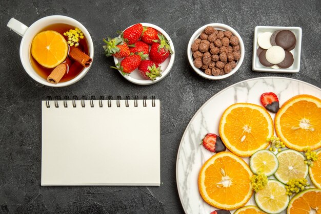 Nahaufnahme von oben eine appetitliche Tasse Tee mit Zitrusfrüchten und Erdbeeren neben den Schalen mit Schokoladenerdbeeren und Haselnüssen weißes Notizbuch und eine Tasse Tee mit Zitrone