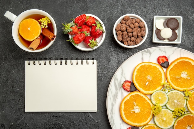 Nahaufnahme von oben eine appetitliche Tasse Tee mit Zitrusfrüchten und Erdbeeren neben den Schalen mit Schokoladenerdbeeren und Haselnüssen weißes Notizbuch und eine Tasse Tee mit Zitrone