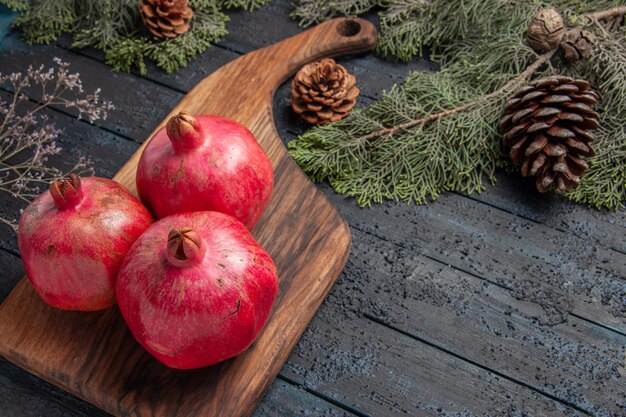 Nahaufnahme von oben reife Granatäpfel rote Granatäpfel auf Schneidebrett neben den Fichtenzweigen mit Zapfen auf grauem Tisch