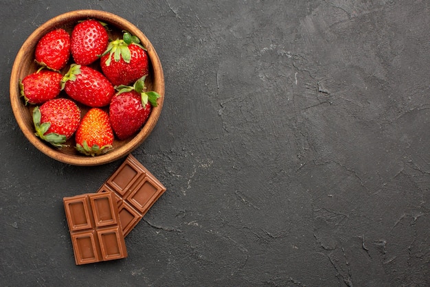 Nahaufnahme von oben Schokolade und Erdbeeren zwei appetitliche Schokoladentafeln neben den Erdbeeren in der Schüssel auf der linken Seite des dunklen Tisches