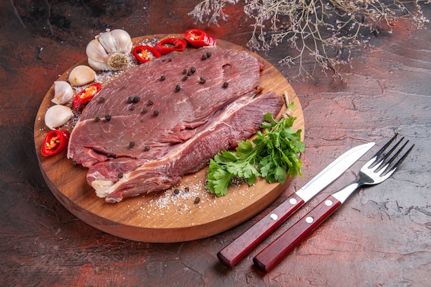 Nahaufnahme von rotem Fleisch auf Holztablett und Knoblauch, grüner Zitronenpfeffer, Zwiebelgabel und Messer auf dunklem Hintergrund