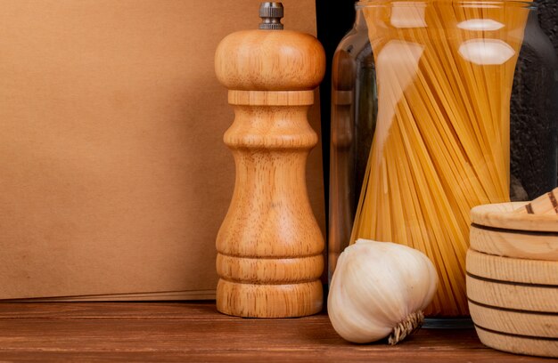 Nahaufnahme von Spaghetti-Nudeln in Glas und Knoblauch mit Salz-Knoblauch-Brecher und Notizblock auf Holzoberfläche und schwarzem Hintergrund mit Kopierraum