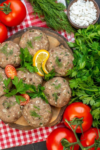 Nahaufnahme von ungekochten Fleischbällchen auf einem Holzbrett auf rotem, abgestreiftem Handtuch und Tomaten auf dunklem Hintergrund