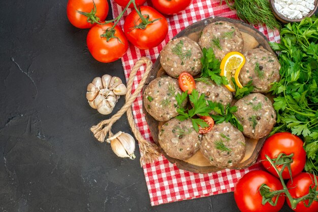 Nahaufnahme von ungekochten Fleischbällchen auf einem Holzbrett auf rotem, abgestreiftem Handtuch und Tomaten-Knoblauch auf der linken Seite auf dunklem Hintergrund