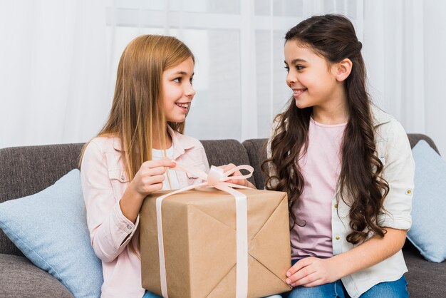 Nahaufnahme von zwei Freundinnen, die auf dem Sofa betrachtet einander beim Öffnen der Geschenkbox sitzen