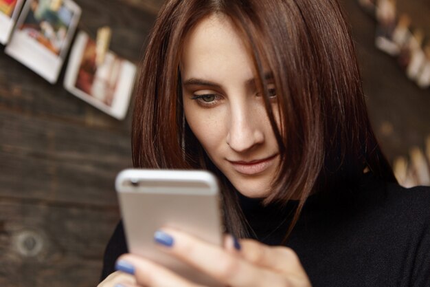 Nahaufnahmeaufnahme der charmanten jungen europäischen Frau, die generisches Handy hält, E-Mail oder Newsfeed über soziale Medien unter Verwendung von Online-Apps prüft.